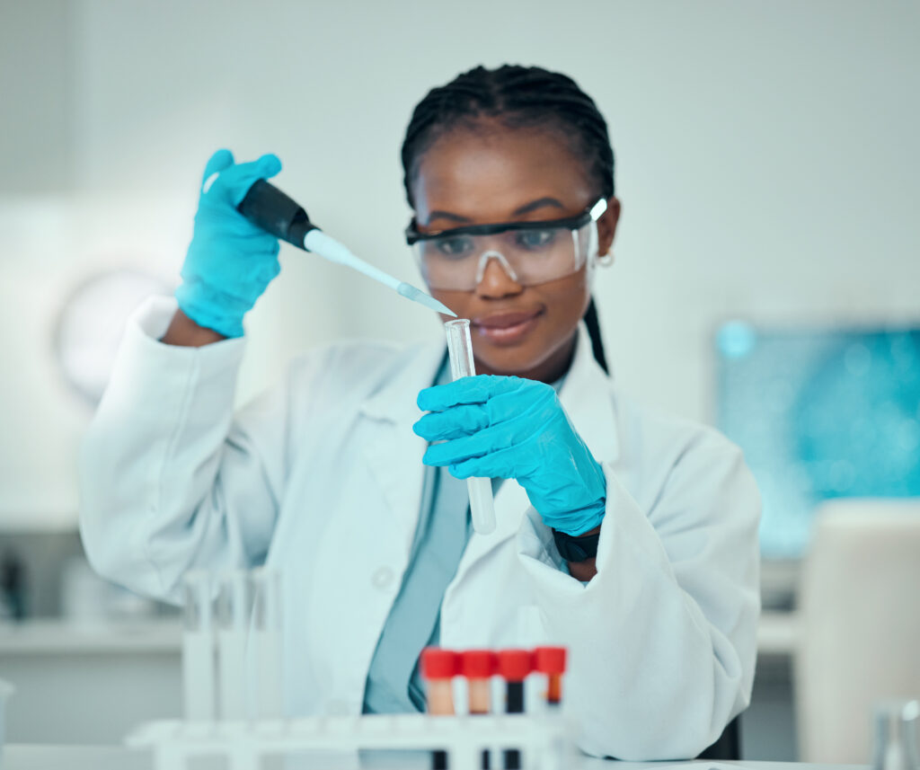 Black woman scientist in a lab coat with safety goggles and blue gloves uses a pipette over a rack of test tubes at a laboratory bench.